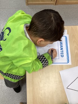 An overhead shot of a child in a yellow safety vest leaning over a wooden table to draw on a paper labeled "WANTED."