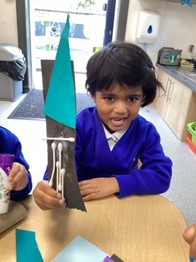A child in a school uniform holds up a finished craft project featuring a green triangle and cotton swabs glued to black paper.