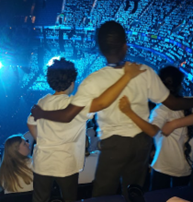 A group of children viewed from behind, wearing white t-shirts and standing with their arms around each other's shoulders while looking toward a blue-lit stage in a large arena.