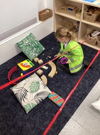 A child in a yellow safety vest sits on a rug inside a play area cordoned off with red tape, investigating coins and blocks with a magnifying glass.