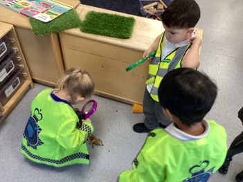 Three children in high-visibility yellow vests use magnifying glasses to examine a small toy animal on the floor of a classroom.