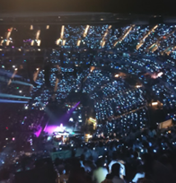 A high-angle, wide view of a packed arena at night, with thousands of audience members holding up glowing blue lights toward a brightly lit stage.
