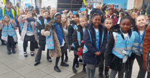 A group of elementary school children standing in a line indoors, wearing blue high-visibility vests and blue lanyards.
