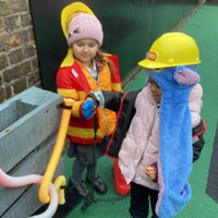 wo children wearing play safety gear and hats interact with colorful, flexible hoses attached to a wooden outdoor structure.