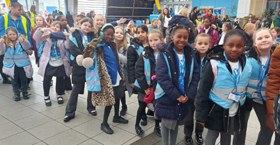 A group of elementary school children standing in a line indoors, wearing blue high-visibility vests and blue lanyards.