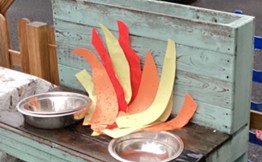 A play fire made from red and yellow paper strips is arranged on a wooden surface next to two metal bowls.