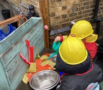 Two children in yellow hard hats and firefighter-themed jackets use a green watering can to pretend to extinguish a paper play fire.