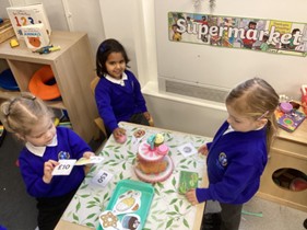 hree young children wearing blue school jumpers sit around a small table in a classroom role-play area labeled "Supermarket." They are interacting with play money, food-themed cards, and a toy cake, practicing social and mathematical skills through play.