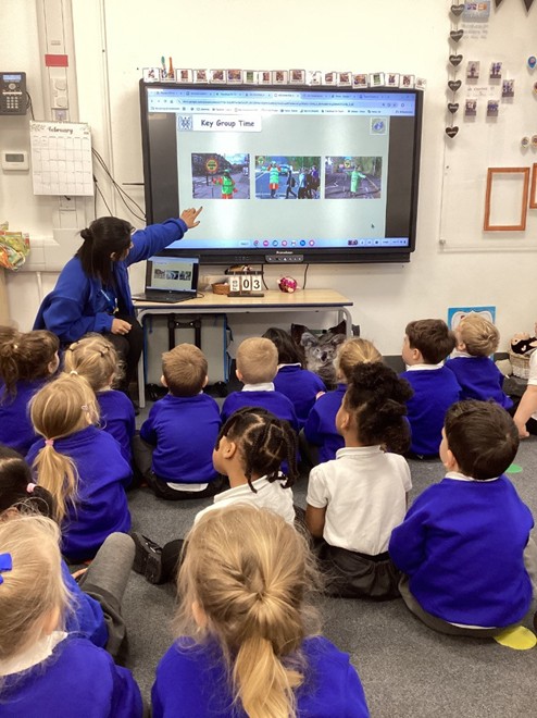 A teacher points to a large interactive screen displaying "Key Group Time" with images of crossing guards, as a group of students watches from the floor.