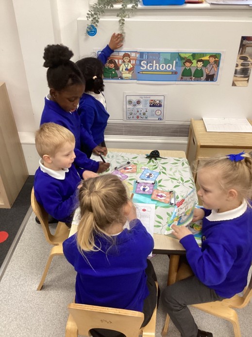 Four young students sit and stand around a small table in a classroom, engaged in a learning activity with colorful cards and books.