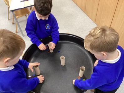 Three children stand around a sensory tray, placing small colored counters into cardboard tubes.