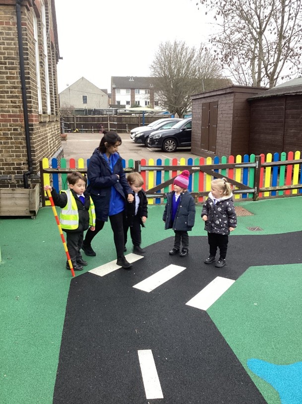 A teacher leads a small group of children across a play-area "road" with white markings, while one student assists by holding a striped crossing pole.