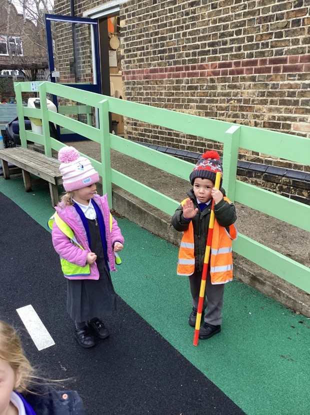 Two children outdoors are practicing road safety; one child wears a high-visibility vest and holds a yellow and red crossing pole while gesturing "stop" with their hand.