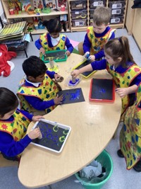 A group of children wearing aprons sit at a table creating art prints using paint and cardboard tubes on black paper.