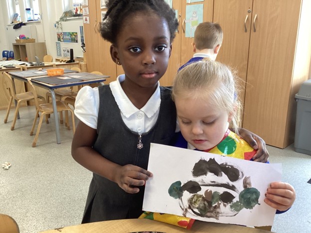 Two young children stand together in a classroom; one child has an arm around the other while they hold up a white sheet of paper with dark paint markings.
