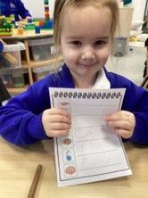 A young girl in a blue school uniform smiles while holding up a worksheet shaped like a notepad. The worksheet features various food icons with handwritten marks or numbers next to them, indicating a completed counting or shopping list activity.