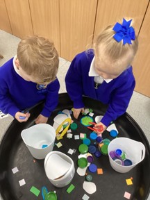 Two children stand at a sensory tray, sorting colorful bottle caps and paper scraps into white buckets.