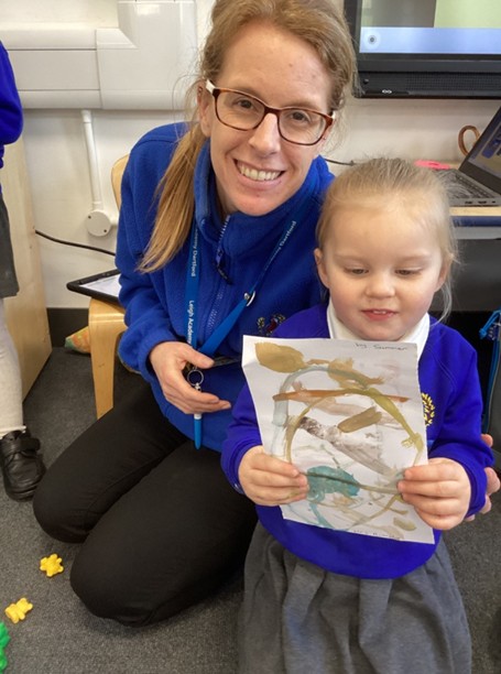 A teacher wearing glasses and a blue fleece smiles next to a young student who is proudly holding up a colorful, abstract finger painting.