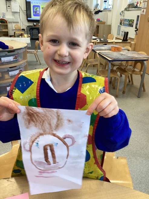 A young boy wearing a yellow protective art smock smiles as he holds up a painting of a face with brown hair and large ears.