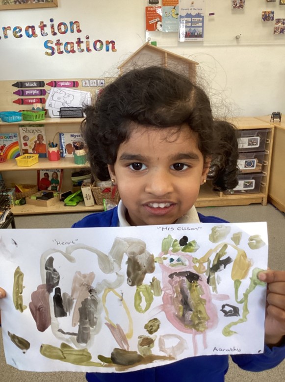A young girl in a classroom stands in front of a "Creation Station" sign, proudly holding up a landscape-oriented finger painting with abstract brown and pink shapes.