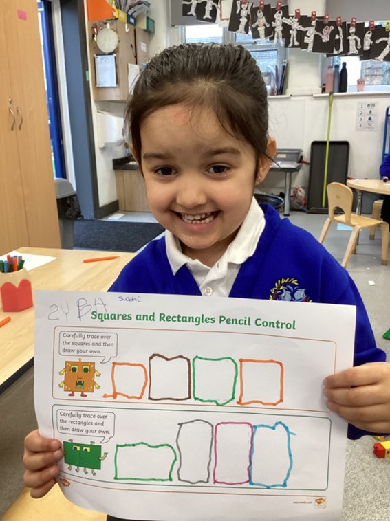 A young girl grins broadly as she displays her completed shape-tracing worksheet, showing squares and rectangles outlined in different colored markers.