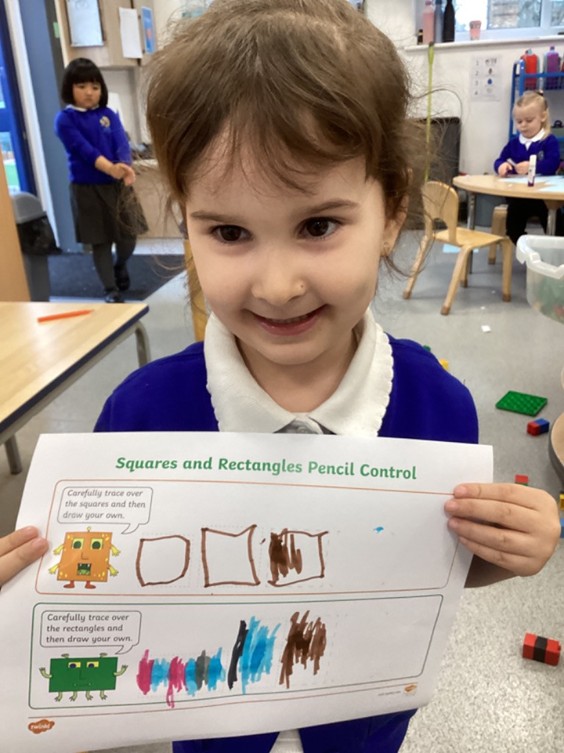 A young girl smiles while holding up a "Squares and Rectangles Pencil Control" worksheet where she has traced and colored in various shapes.