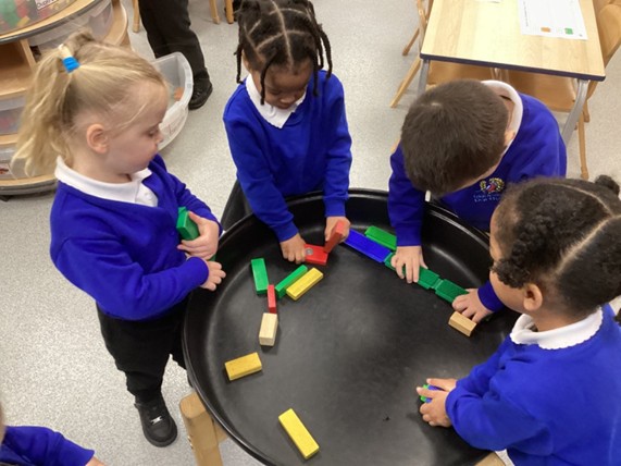 Four young children in blue school jumpers stand around a large black tray, collaborating to arrange colorful rectangular building blocks.