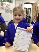 young girl in a blue school jumper proudly displays a "shopping list" worksheet in a classroom setting. The sheet shows images of fruit and bread with her handwritten attempts at letters and numbers next to each item