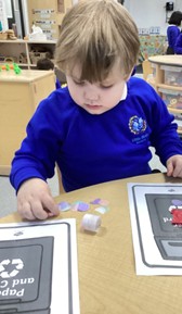 A child sits at a desk carefully pasting small paper shapes onto a worksheet featuring a recycling bin.