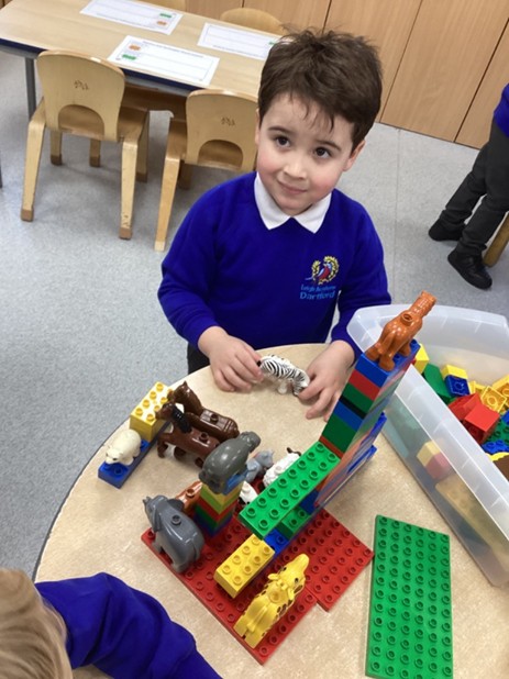 A young boy in a blue jumper plays at a table with colorful building blocks and plastic animal figures, including a zebra and a polar bear.