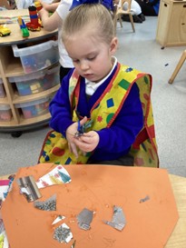 A young girl wearing a yellow patterned apron over her blue school jumper carefully uses scissors to cut small pieces of paper. Several cut-out scraps are scattered across a large orange sheet of paper on the table in front of her.