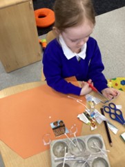 A young girl with her hair in a ponytail works at a table, using red safety scissors to cut out images from a magazine. A muffin tin nearby holds cotton swabs, and various paper scraps are scattered on a large orange sheet of craft paper.