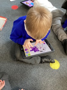 A young child in a blue jumper sits on a grey carpeted floor using a tablet. The screen shows a digital coloring activity of a dinosaur being colored purple.