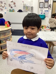 A young child holds up a drawing of a truck colored in blue and red.