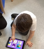 A young boy in a white polo shirt leans over a tablet on the floor, using his finger to interact with a grid of colorful digital images.