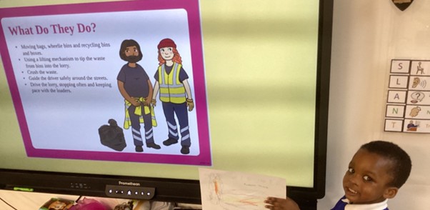 A young student stands next to a large screen displaying information about refuse collectors, smiling while holding up a hand-drawn picture.