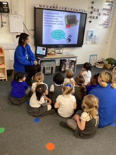 A group of children sit on a carpeted floor in front of a large interactive whiteboard. A teacher in a blue cardigan sits to the side as they look at a story on the screen about a penguin receiving a brand new tablet.