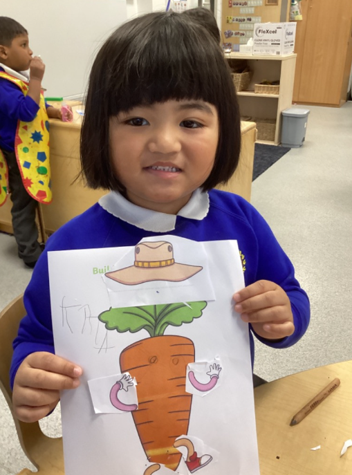 A young girl in a blue jumper holds up a "Build a Vegetable Character" worksheet where she has glued a hat and arms onto a large carrot illustration.