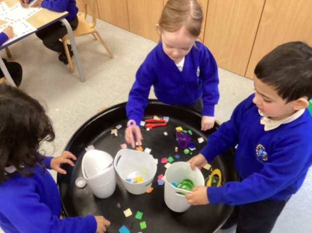 Three children gather around a black sensory tray, using tongs to pick up and sort play materials into containers.