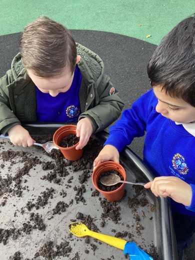 Two students in blue school uniforms using spoons to scoop soil into small plant pots.