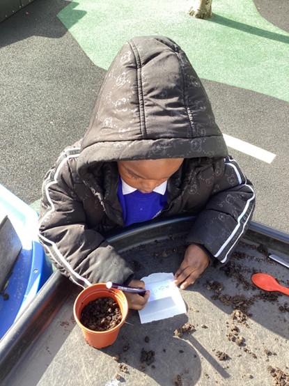 A student wearing a winter coat writing on a small white label next to a plant pot filled with soil.