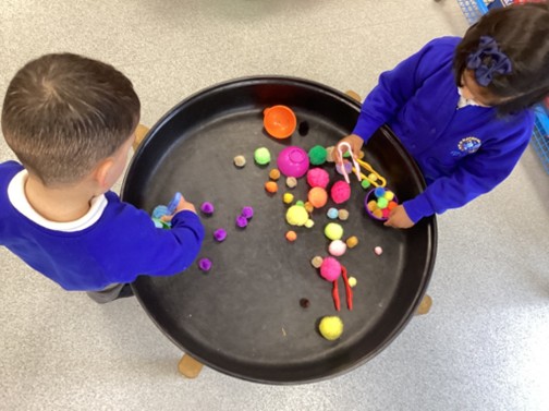 A top-down view of two students using tongs and small bowls to play with colorful pom-poms inside a large black tray.