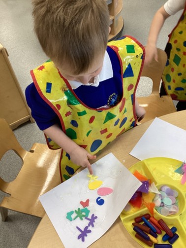 A student wearing a yellow craft apron working on a collage with small, colorful shapes, next to a tray of craft supplies.