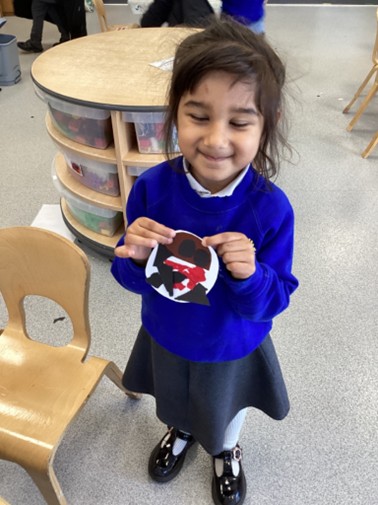 A young girl in a blue school sweater smiles while holding up a circular paper craft decorated with red and black shapes.