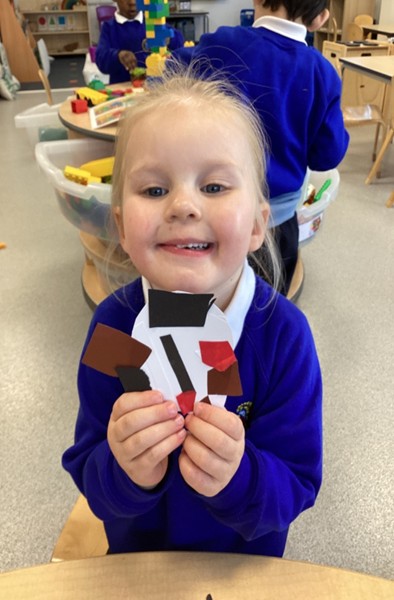 A smiling young girl holds up an oval-shaped paper craft featuring glued-on brown, red, and black paper segments.