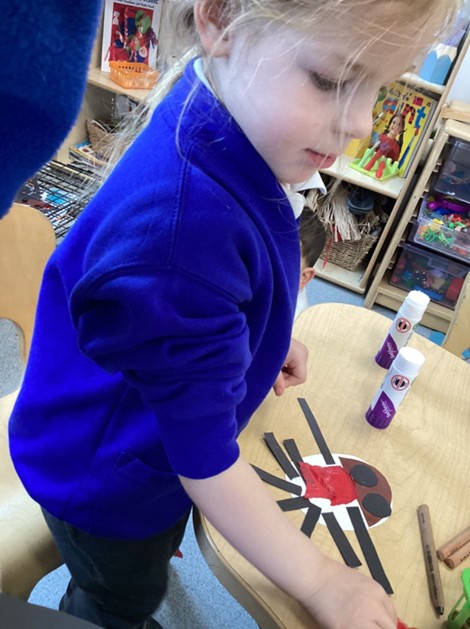 young girl with blonde hair works at a table, carefully gluing red tissue paper and black strips onto a white paper base.