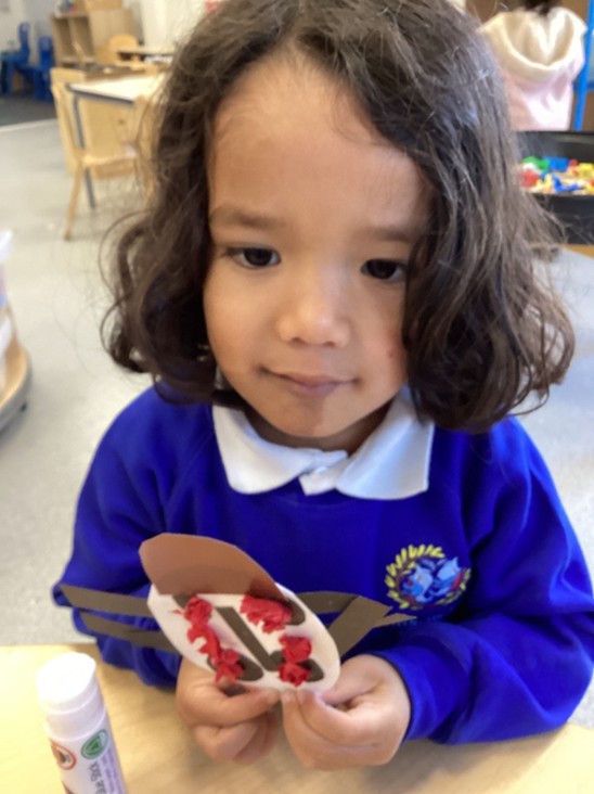 A young girl with dark curly hair looks down at her completed craft, which features red crumpled paper and black paper "legs."