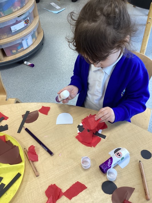 A high-angle shot of a young girl sitting at a table, using a glue stick to attach red tissue paper to her craft project.