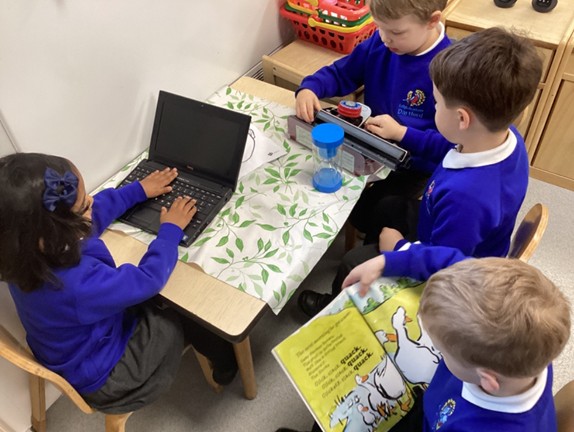Three students gathered around a table; one uses a laptop, one plays with a toy cash register, and another holds open a picture book showing illustrations of ducks.