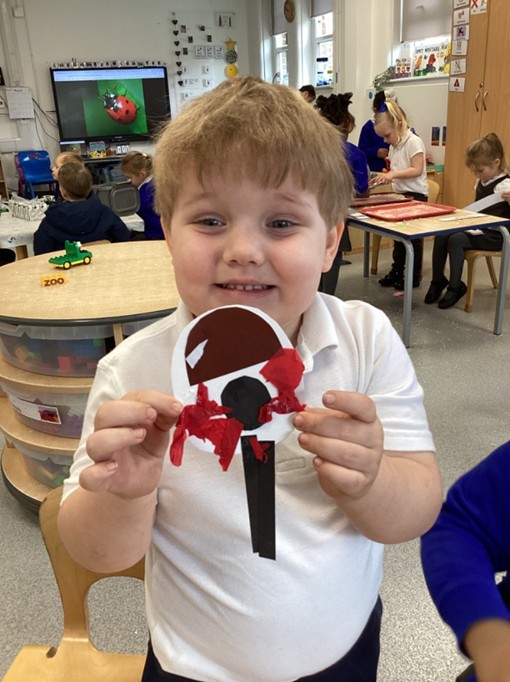A young boy in a white polo shirt smiles broadly while displaying his finished paper craft; in the background, a screen shows a ladybird.